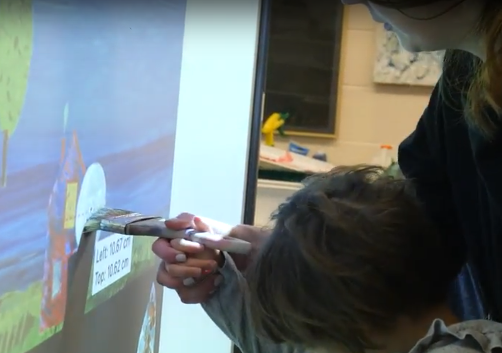 Caregiver is helping a student to hold a brush and move it on a screen.